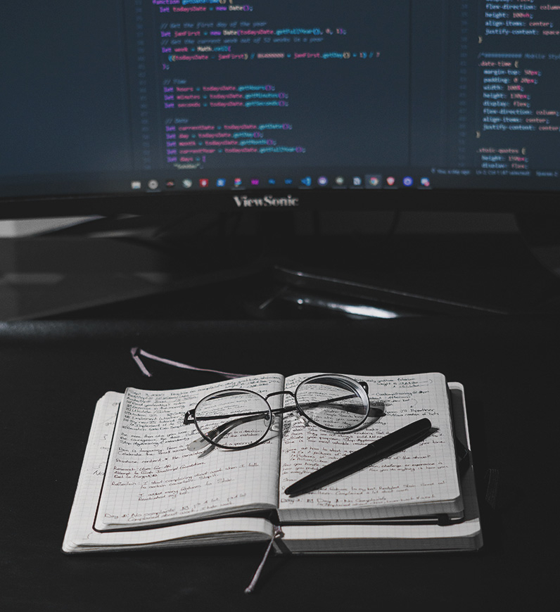 Coding notes next to a notebook, glasses, and a computer monitor. High-quality web development is essential for business success.